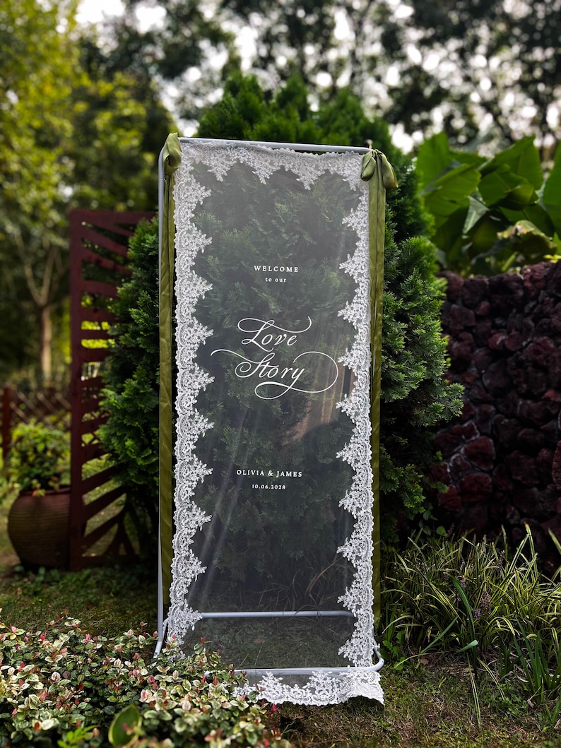 May include: A rectangular vertical sign featuring a clear acrylic panel framed in white metal. The panel is draped with white lace trim along the sides and includes the white printed text: WELCOME to our Love Story OLIVIA & JAMES 10.04.2028.
