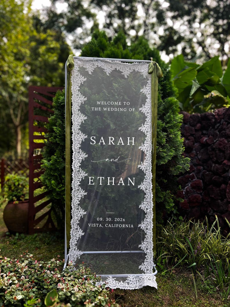 May include: A rectangular clear acrylic wedding sign held in a white metal frame, draped with white lace trim along the vertical edges and olive green ribbons at the top. The text reads: WELCOME TO THE WEDDING OF SARAH and ETHAN 09. 30. 202x VISTA, CALIFORNIA.