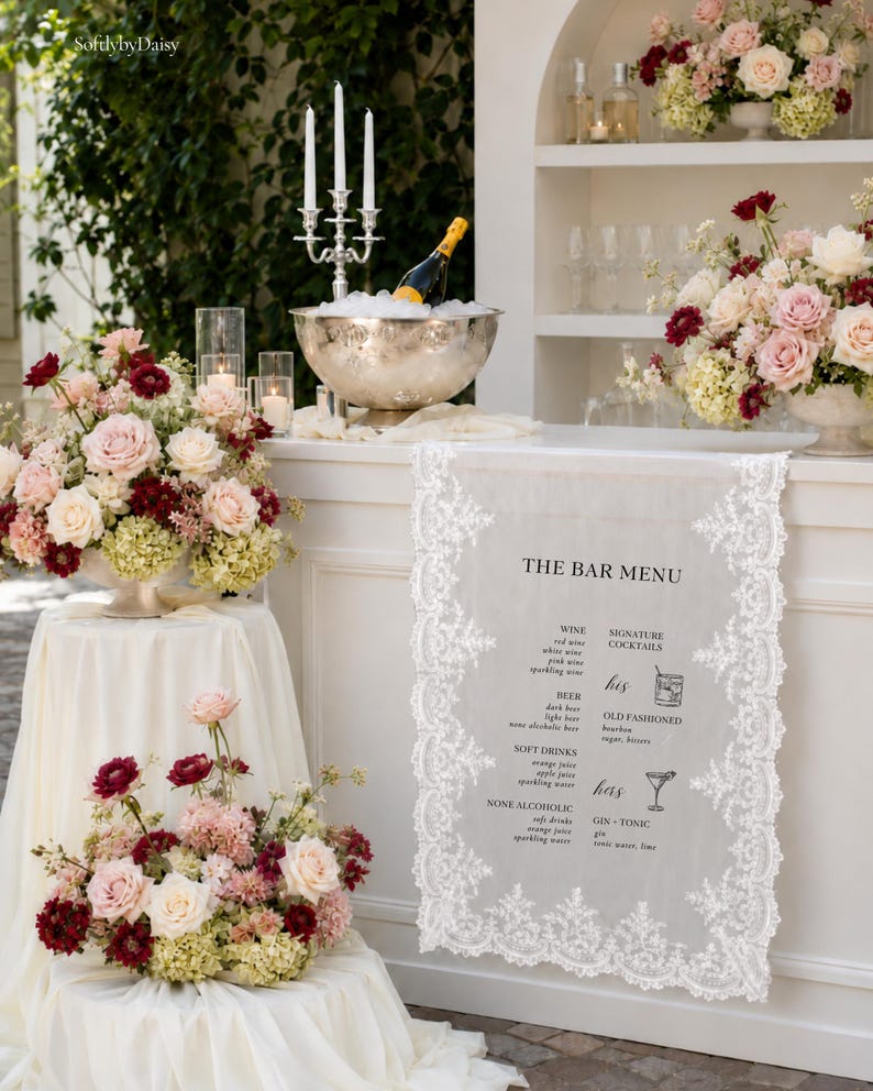 以下が含まれることがあります： 複雑なレースの縁取りが施された白い布製のバーメニュー看板。ワイン、ビール、ソフトドリンク、オールドファッションやジントニックなどのシグネチャーカクテルが黒い文字で記載されており、花で飾られた白いバーカウンターの上に展示されています。