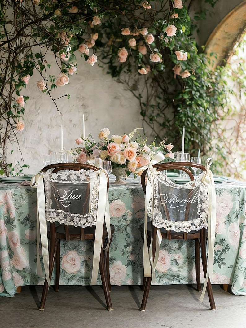 May include: Wedding reception setting with two wooden chairs adorned with lace and ribbon. One chair has a sign that says "Just" and the other says "Married". A floral arrangement and table setting are visible.