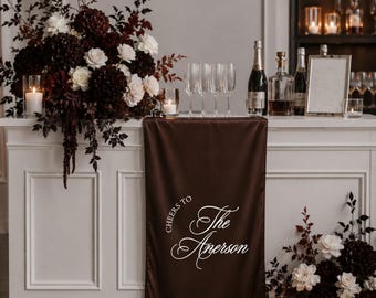Puede incluir: Un camino de mesa de satén marrón oscuro drapeado sobre una barra blanca, con el texto blanco impreso "CHEERS TO The Amerson" en una mezcla de fuentes serif y script. El camino está acentuado por arreglos florales oscuros y portavelas de cristal.