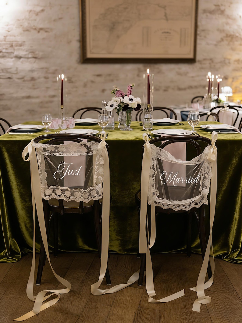 May include: Wedding reception table setting with a green velvet tablecloth, candles, and floral arrangements. Two chairs are decorated with lace and ribbons, with the words "Just" and "Married" displayed.