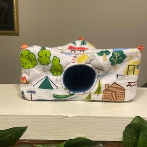 May include: A white, camera-shaped pet bed with a dark blue opening. The bed is decorated with green trees, a campfire, a tent, and a cabin. The word "CAMP" is printed on the front.