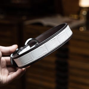 Könnte beinhalten: Ein weißer Ledergürtel mit braunem Lederuntergrund. Der Gürtel ist aus Krokodilleder und hat eine silberne Schnalle.