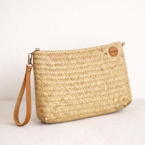 May include: A rectangular woven straw clutch with a tan leather wrist strap. The bag has a small, round leather patch with the text "cclc" on it. The bag is made of natural-colored straw and has a zipper closure.