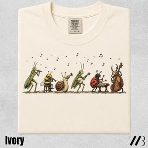 Puede incluir: Una camiseta de color marfil con una ilustración gráfica de seis insectos tocando instrumentos musicales, incluyendo un saltamontes con flauta, una abeja con clarinete, un caracol con trompeta, un saltamontes con banjo, una mariquita con batería y un escarabajo con contrabajo.