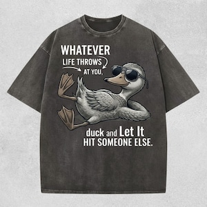 Pode incluir: Camiseta cinza escuro com um pato de desenho animado usando óculos de sol. O texto diz: "WHATEVER LIFE THROWS AT YOU, duck and Let It HIT SOMEONE ELSE." O pato é cinza claro com bico amarelo e óculos de sol pretos.