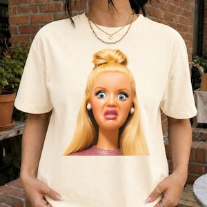 Puede incluir: Camiseta color crema con un primer plano de la cara de una muñeca con grandes ojos azules y una expresión de sorpresa. La muñeca tiene el pelo rubio y lleva una camiseta rosa. La camiseta es de estilo casual.