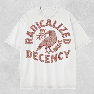 Puede incluir: Camiseta blanca con cuello redondo y mangas cortas. La camiseta presenta un diseño circular en marrón rojizo con las palabras "RADICALIZED BY BASIC DECENCY" que rodean una ilustración detallada de un cuervo sosteniendo una ramita.