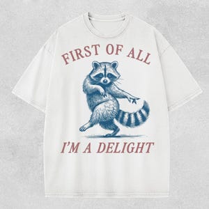 Puede incluir: Camiseta blanca con una ilustración azul de un mapache bailando. El texto "FIRST OF ALL" está encima del mapache, y "I'M A DELIGHT" debajo, en color burdeos.