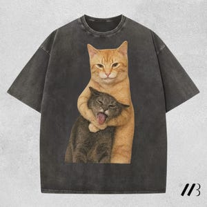 Op de afbeelding: Donkergrijs t-shirt met een afbeelding van twee katten. Een oranje tabby kat omhelst een grijze kat, die zijn tong uitsteekt. Het shirt heeft een vintage, gewassen uitstraling en een relaxte pasvorm.