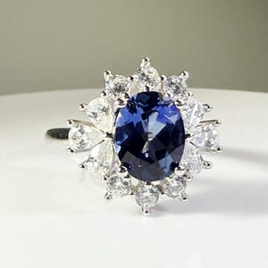 Könnte beinhalten: Ein silberner Ring mit einem großen, oval geschliffenen, tiefblauen Edelstein, umgeben von kleineren, klaren Edelsteinen. Die klaren Steine sind in einem floralen oder sternförmigen Muster um den zentralen Stein angeordnet und erzeugen einen funkelnden Effekt. Der Ring liegt auf einer weißen Oberfläche.