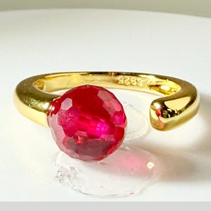 May include: A gold-colored ring with a large, faceted, red gemstone. The ring has an open design, with the gemstone prominently displayed. The band is smooth and shiny, and the ring appears to be made of metal.