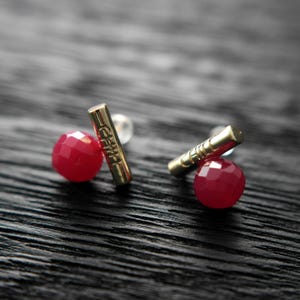 Puede incluir: Un par de pendientes dorados con un diseño de barra y una cuenta roja redonda y facetada. La barra tiene texto grabado. Los pendientes están sobre una superficie oscura y texturizada.
