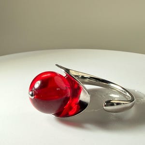 May include: A silver bracelet with a large, round, translucent red bead. The bead is set in a silver metal cup, with a curved, open design. The bracelet has a modern, artistic style.