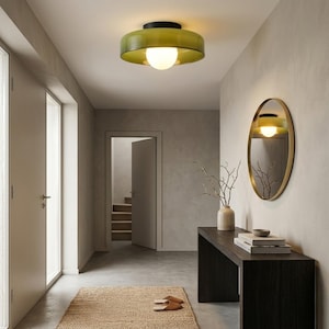 May include: A hallway featuring a green and black ceiling light with a spherical bulb. An oval mirror with a gold frame is on the wall. A dark wood console table holds a vase and books. A woven rug and slippers are on the floor.