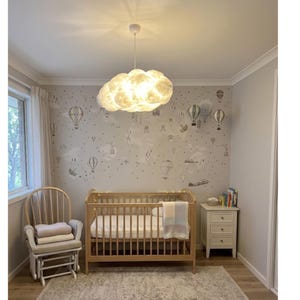 May include: A nursery with a light wood crib, a rocking chair, and a white nightstand. The walls are decorated with a hot air balloon pattern. A cloud-shaped light fixture hangs from the ceiling, illuminating the room. A soft blanket rests on the crib.