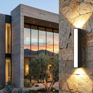 Pode incluir: Design arquitetônico moderno com um edifício de concreto e grandes janelas de vidro refletindo um pôr do sol. Uma luminária externa retangular preta é montada em uma parede de pedra, projetando a luz para baixo. Uma árvore está em primeiro plano.