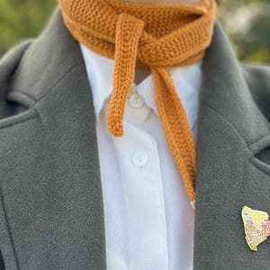 Pode incluir: Cachecol de malha laranja amarrado sobre uma camisa branca e um casaco verde escuro. Um broche dourado com um design colorido é preso ao casaco. O cachecol tem um tom quente, criando um contraste com as outras peças de roupa.