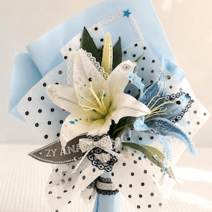 May include: A bouquet of white and blue lilies decorated with silver glitter, small blue star accents, and rhinestones. The flowers are wrapped in light blue paper and white polka-dot tissue paper, featuring a black lace bow and a tag reading ZY ANA.
