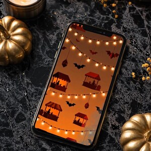 May include: A smartphone displays a Halloween-themed wallpaper with bats, string lights, and market stalls against an orange gradient. Gold pumpkins and lit candles surround the phone on a black marble surface, creating a festive autumn scene.
