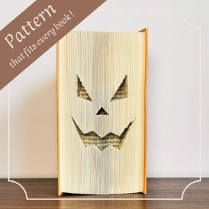 Könnte beinhalten: Ein Buch mit orangefarbenen Einbänden und Seiten, die so geschnitzt sind, dass sie einem Kürbisgesicht ähneln. Der Text "Pattern that fits every book!" wird diagonal in einem braunen Banner angezeigt.