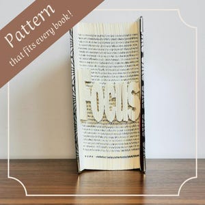 Könnte beinhalten: Ein Buch mit dem Wort "FOCUS" in die Seiten geschnitten. Das Buch steht auf einer Holzoberfläche. Der Buchrücken hat ein schwarz-weißes Design. Text in einem braunen Banner lautet "Pattern that fits every book!"