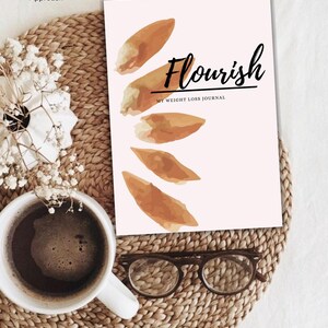 Könnte beinhalten: Ein rosa-weißes Journal mit dem Titel "Flourish: My Weight Loss Journal" in schwarzer Schrift. Der Einband des Journals zeigt ein Aquarelldesign mit vier braunen Blättern.