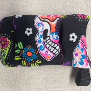Puede incluir: Una bolsa de tela negra con cierre de cremallera y una correa negra. La bolsa está decorada con un diseño de calavera de azúcar colorido y patrones florales.
