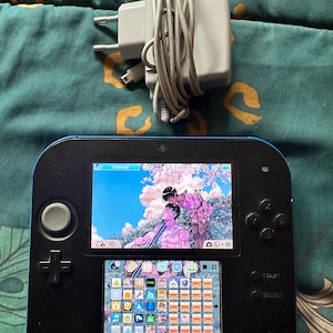 May include: A black and blue handheld game console with a dual-screen display showing an anime-style illustration on the top screen and a grid of application icons on the bottom screen, accompanied by a grey power adapter with a coiled cable.