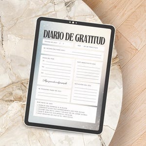 May include: A digital tablet displaying a gratitude journal titled "DIARIO DE GRATITUD" in Spanish. The journal features sections for daily intentions, gratitude, and reflections. The tablet is on a marble-like surface.