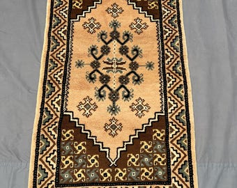 Puede incluir: Una alfombra rectangular con un centro color melocotón y un borde detallado. El borde presenta un patrón geométrico repetitivo en marrón, negro y verde. La alfombra tiene flecos en ambos extremos, agregando un toque decorativo.
