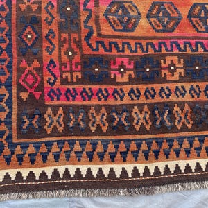 May include: Close-up of a section of a patterned rug. The rug features a complex design with geometric shapes in shades of brown, orange, blue, and pink. The edges have a fringe detail. The rug's design is intricate and detailed.