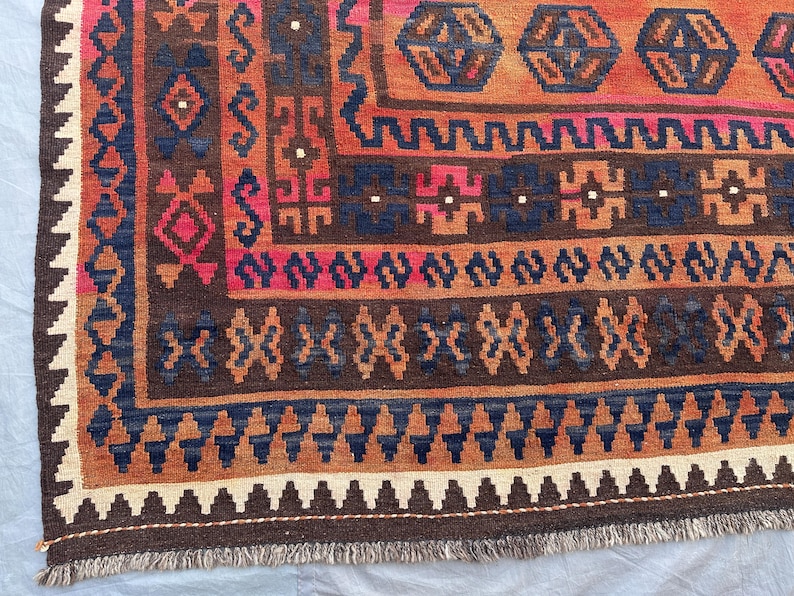 May include: Close-up of a section of a patterned rug. The rug features a complex design with geometric shapes in shades of brown, orange, blue, and pink. The edges have a fringe detail. The rug's design is intricate and detailed.