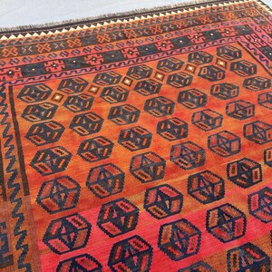 May include: A rectangular, hand-woven rug with a geometric pattern. The rug features a repeating design of dark blue and brown geometric shapes on a red and orange background. The border has a complex pattern with shades of brown, red, and black.