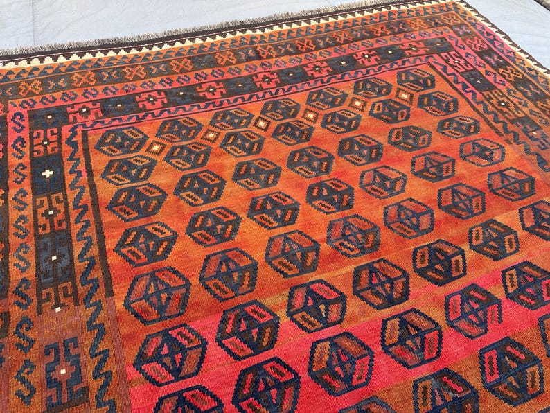 May include: A rectangular, hand-woven rug with a geometric pattern. The rug features a repeating design of dark blue and brown geometric shapes on a red and orange background. The border has a complex pattern with shades of brown, red, and black.