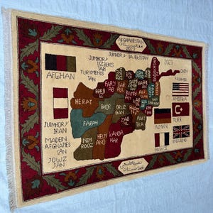 May include: A rectangular area rug featuring a map of Afghanistan with labeled provinces in various colors. The border is deep red with green floral patterns. It includes flags of several nations, the text "AFGHANISTAN 2023", and "MADE IN AFGHANISTAN".