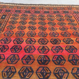 May include: A rectangular rug with a geometric pattern. The rug features a repeating pattern of dark blue and brown geometric shapes on a red and orange background. The border is a darker brown with geometric designs.