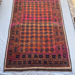 May include: A rectangular, hand-woven rug with a geometric pattern. The rug features a central field of orange and pink hues, surrounded by a border of dark brown, navy blue, and white. The design includes repeating hexagonal shapes and intricate border details.