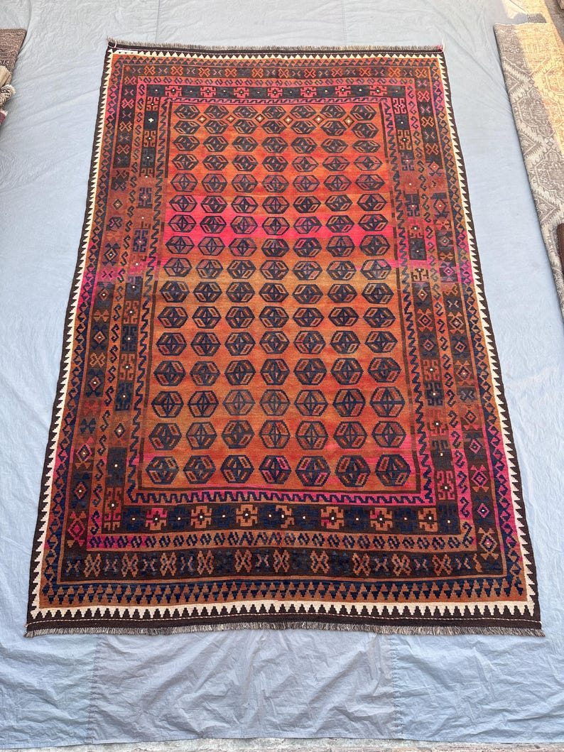 May include: A rectangular, hand-woven rug with a geometric pattern. The rug features a central field of orange and pink hues, surrounded by a border of dark brown, navy blue, and white. The design includes repeating hexagonal shapes and intricate border details.
