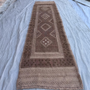 May include: A rectangular brown rug with a central diamond pattern and a border of repeating geometric shapes. The rug has a fringe on both ends and is displayed on a light-colored surface.