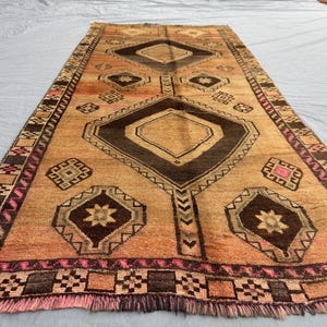 Könnte beinhalten: Ein rechteckiger Teppich mit einem geometrischen Muster in Braun-, Beige- und Rosatönen. Der Teppich hat Diamanten- und Quadratformen mit einem Fransenrand. Das Design ist symmetrisch und schafft eine traditionelle Ästhetik.