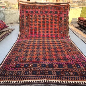 May include: A large, rectangular rug with a geometric pattern in shades of red, orange, and brown. The rug features a central design of repeating octagons and a border with intricate detailing. The rug is displayed outdoors, with other rugs visible in the background.