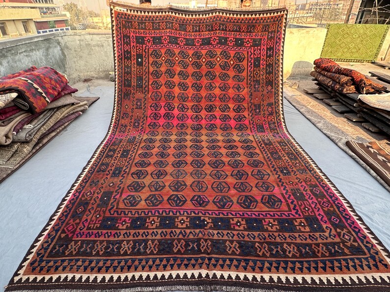May include: A large, rectangular rug with a geometric pattern in shades of red, orange, and brown. The rug features a central design of repeating octagons and a border with intricate detailing. The rug is displayed outdoors, with other rugs visible in the background.