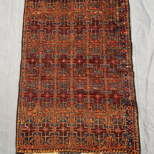 May include: A rectangular area rug featuring a repeating grid pattern of dark blue and deep red cross-like motifs outlined in orange. The rug has a dark border with small orange accents and short, light-coloured fringe along the top and bottom edges.