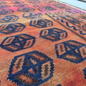 May include: Close-up of a hand-woven rug featuring a repeating pattern of blue and brown geometric shapes on a reddish-orange background. The rug has a border with a zig-zag design and a pink section.