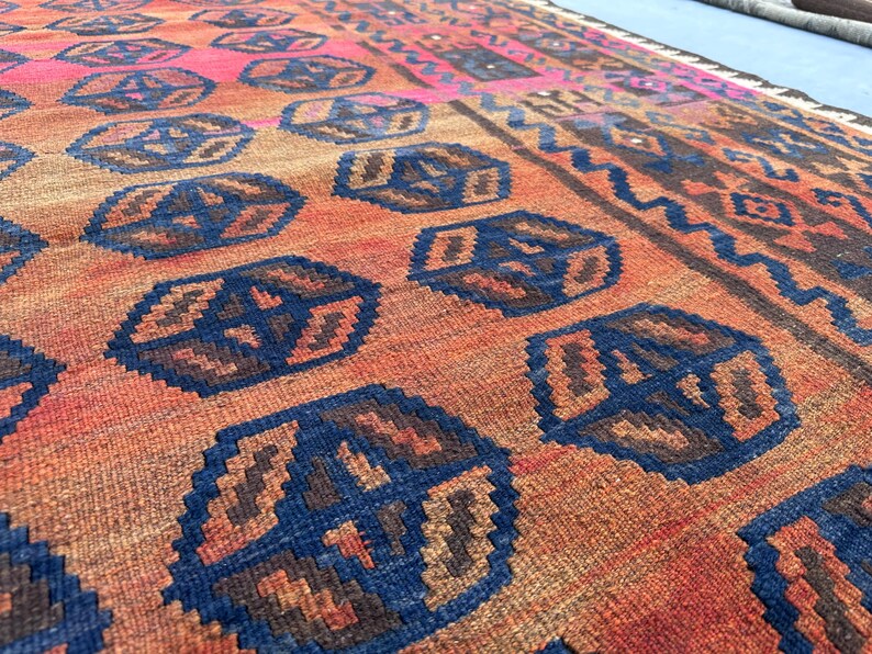 May include: Close-up of a hand-woven rug featuring a repeating pattern of blue and brown geometric shapes on a reddish-orange background. The rug has a border with a zig-zag design and a pink section.