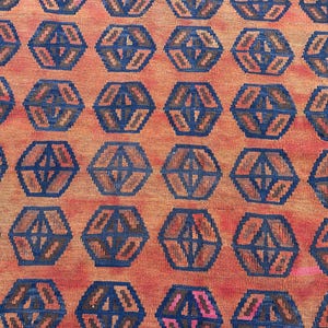 May include: Close-up of a woven rug with a repeating geometric pattern. The rug has a warm orange background with dark blue and brown hexagonal shapes. The design creates a textured, patterned surface.