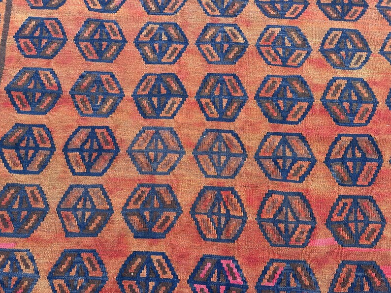 May include: Close-up of a woven rug with a repeating geometric pattern. The rug has a warm orange background with dark blue and brown hexagonal shapes. The design creates a textured, patterned surface.