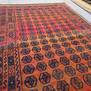 May include: A large, rectangular rug with a geometric pattern. The rug features a gradient of colours, transitioning from a vibrant pink to orange and brown. Dark blue geometric shapes are evenly spaced across the rug's surface. The border has intricate patterns in various colours.